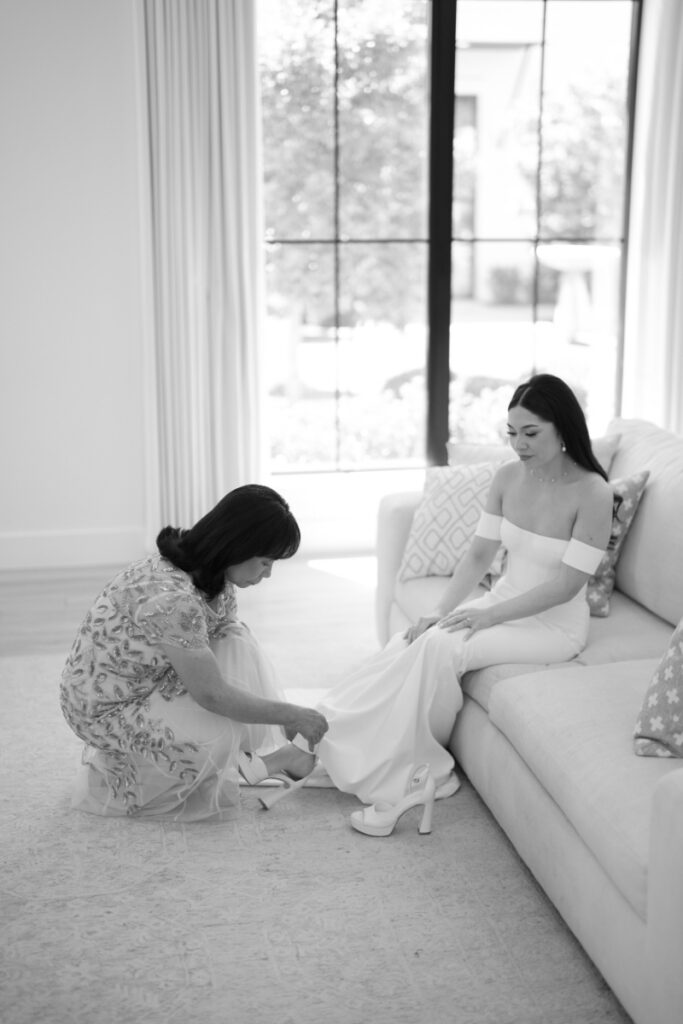 A woman helps another woman in a white dress put on her shoe as she sits on a couch in a bright room.