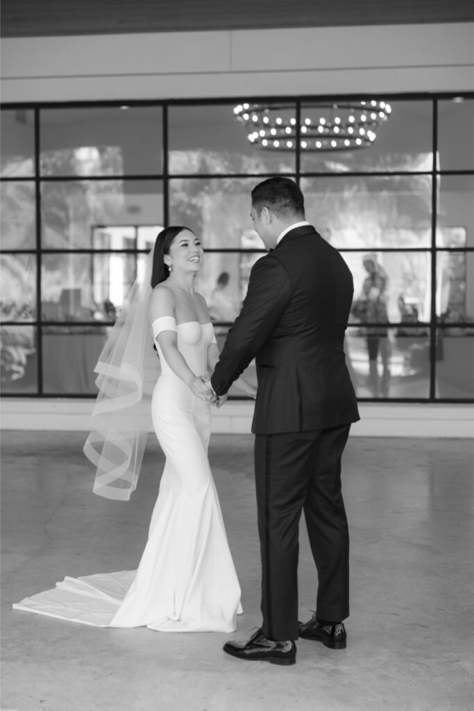 A bride in a strapless gown and veil holds hands with a groom in a dark suit, both smiling, standing inside The Arlo Austin in front of large windows.