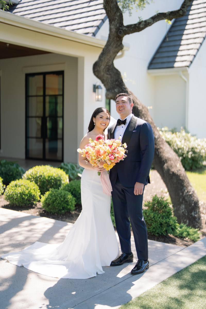 A bride in a white dress and a groom in a dark suit stand outdoors in front of a house, smiling and holding a bouquet of flowers.