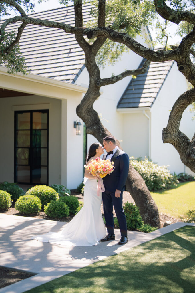 A bride and groom stand together outside The Arlo Austin, with the bride holding a large bouquet of flowers under a tree.