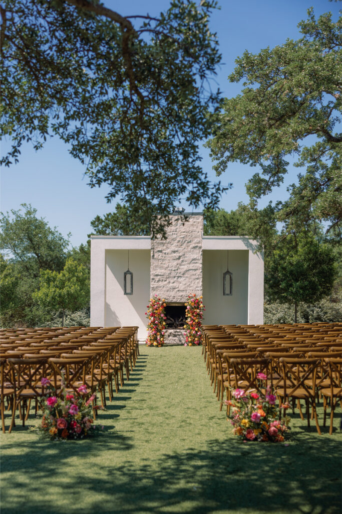 Outdoor wedding ceremony setup with rows of wooden chairs facing a white altar adorned with colorful floral arrangements, surrounded by greenery and trees under a clear blue sky.