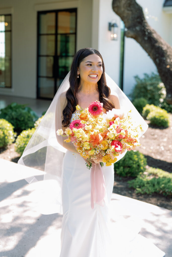 A bride in a white dress and veil holds a large bouquet of colorful flowers while standing outdoors near a modern building.