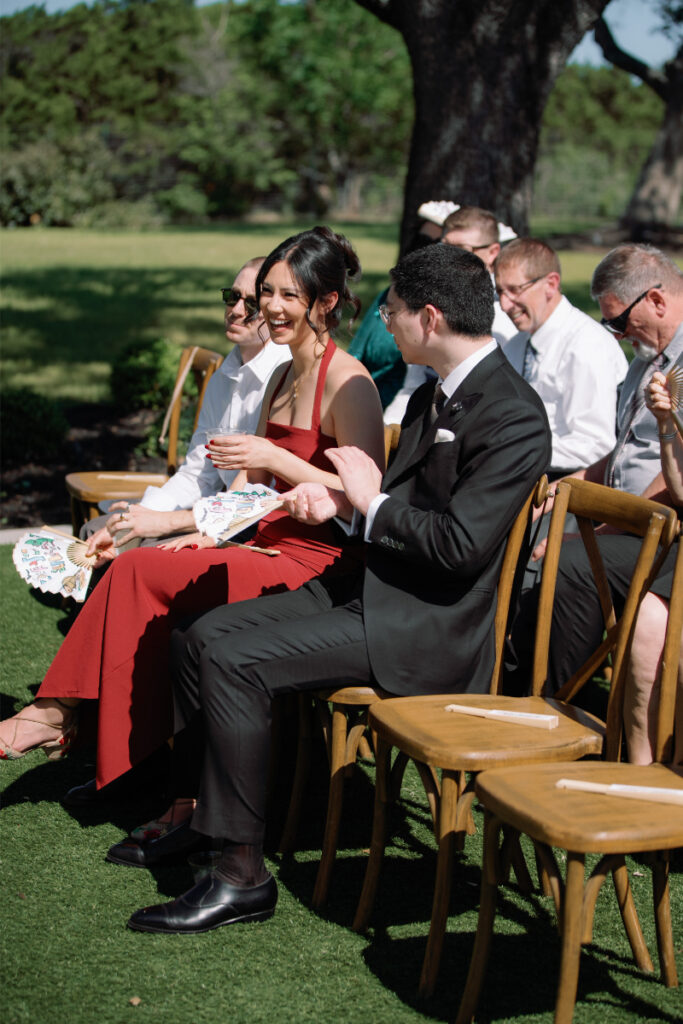 A group of people sit on wooden chairs outdoors at a formal event, dressed in semi-formal attire, with some holding paper fans in sunny weather.