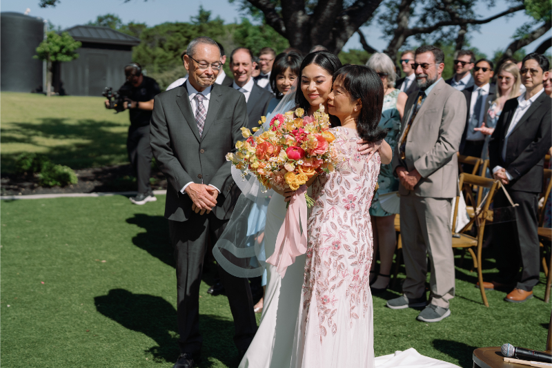 A bride holding a bouquet hugs a woman in a pink dress outdoors, while an older man and wedding guests look on during a ceremony.
