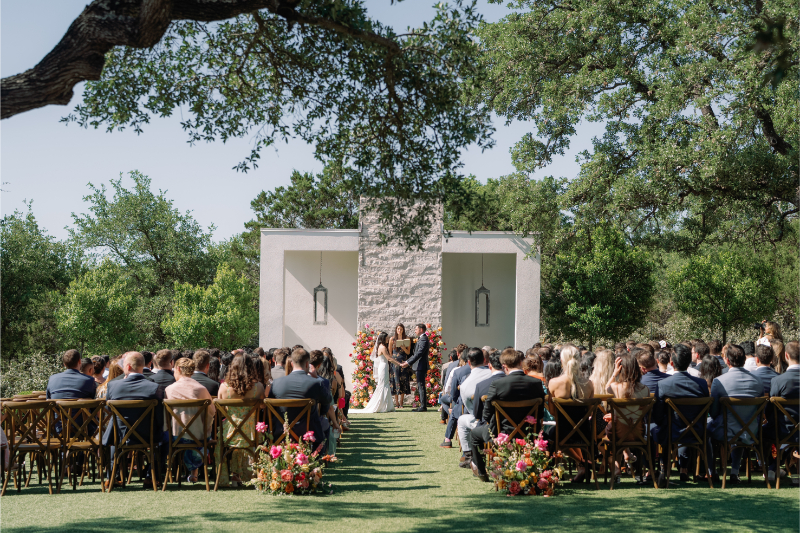 A bride and groom stand at an outdoor altar at The Arlo Austin with guests seated on both sides, surrounded by greenery and floral arrangements on a sunny day.