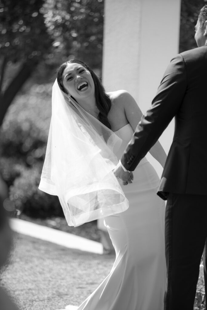A bride in a strapless dress and veil laughs while holding hands with a person in a suit during an outdoor wedding ceremony.
