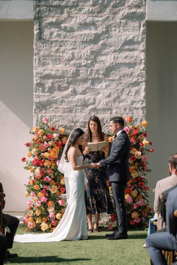 A bride and groom stand facing each other, holding hands, during an outdoor wedding ceremony at The Arlo Austin with a floral backdrop and an officiant reading from a book.