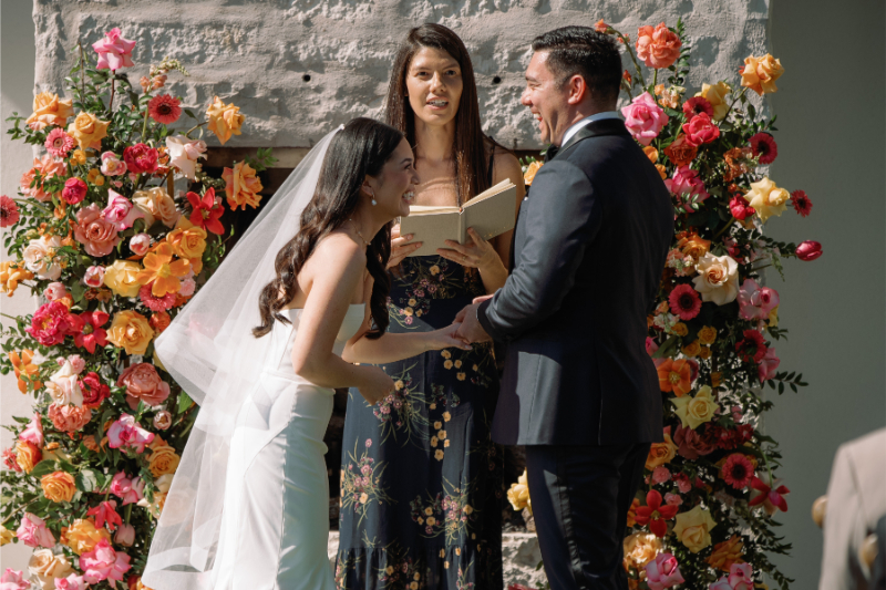 A bride and groom stand laughing and holding hands during their wedding ceremony, with an officiant and a colorful floral backdrop behind them.