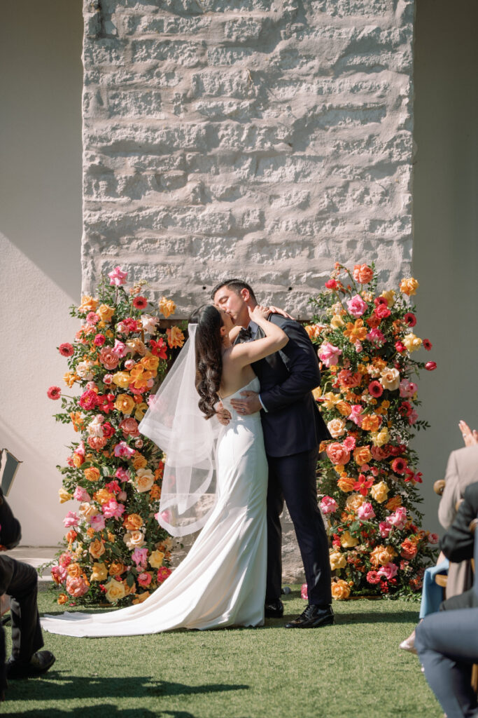 A bride and groom share a kiss in front of a floral arrangement and stone wall during their outdoor wedding ceremony.
