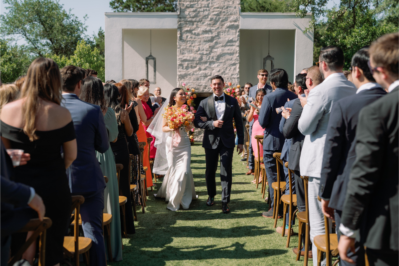 A bride and groom walk down the aisle together outdoors, surrounded by seated guests clapping and standing on both sides.