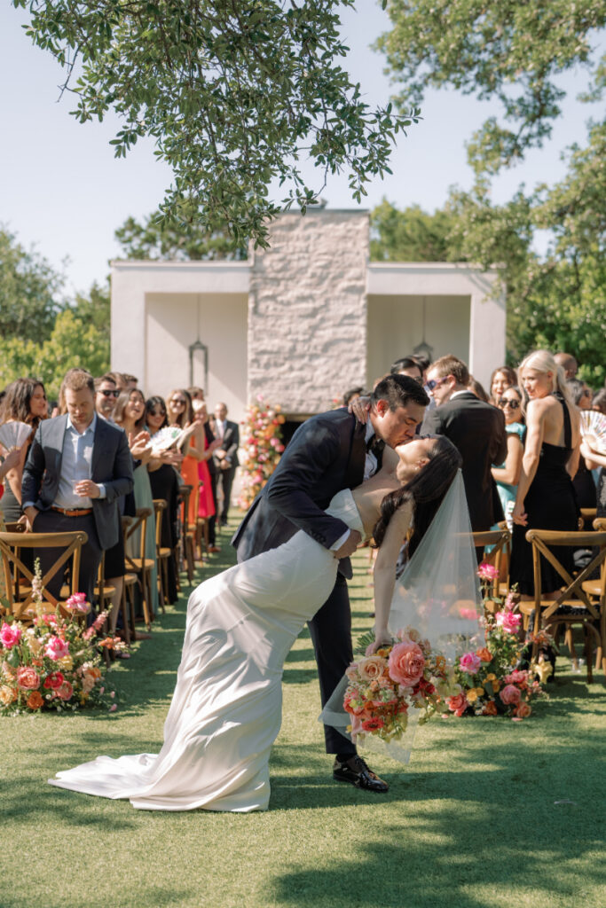 A bride and groom kiss during an outdoor wedding ceremony at The Arlo Austin, surrounded by guests and vibrant floral arrangements on a sunny day.