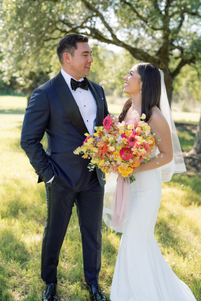 A bride in a white dress holding a colorful bouquet stands beside a groom in a dark suit outdoors under trees, both smiling at each other.