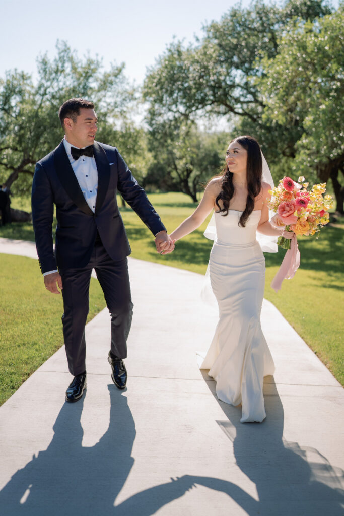 A bride in a white dress holds a bouquet and walks hand-in-hand with a groom in a navy tuxedo on a sunny path with trees in the background.