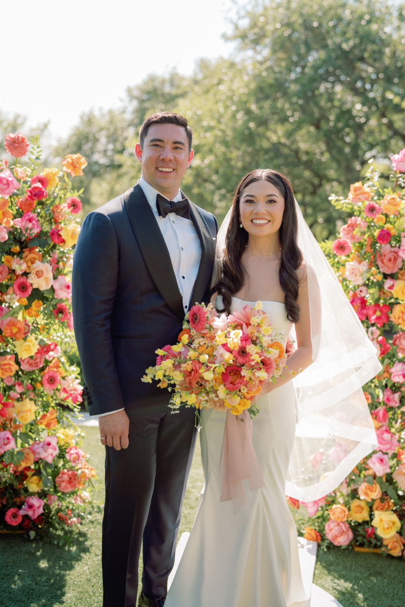 A bride and groom stand outdoors, smiling, surrounded by vibrant floral arrangements. The bride holds a colorful bouquet and wears a white dress and veil; the groom wears a black tuxedo.