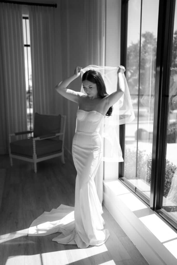 A woman in a fitted wedding dress adjusts her veil while standing by a large window in a sunlit room.