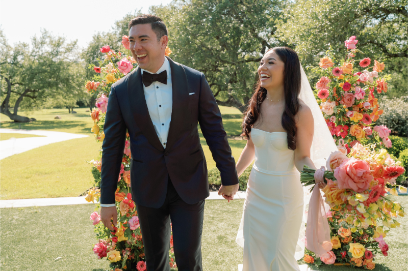 A bride and groom, holding hands and smiling, stand outdoors in front of colorful floral arrangements on a sunny day.