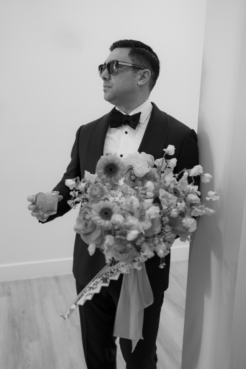A man in a tuxedo and sunglasses holds a large bouquet of flowers in one hand and a drink in the other, standing indoors against a wall.
