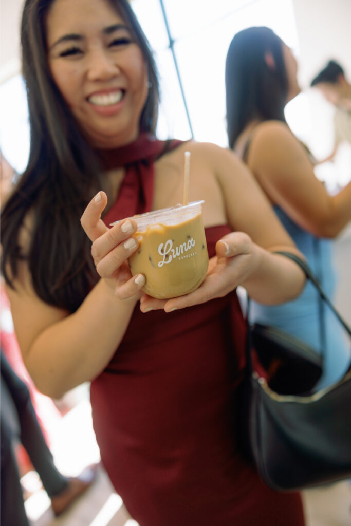 A woman in a sleeveless maroon dress smiles while holding an iced drink in a clear cup labeled "Luna" with a straw. Other people are visible in the background.