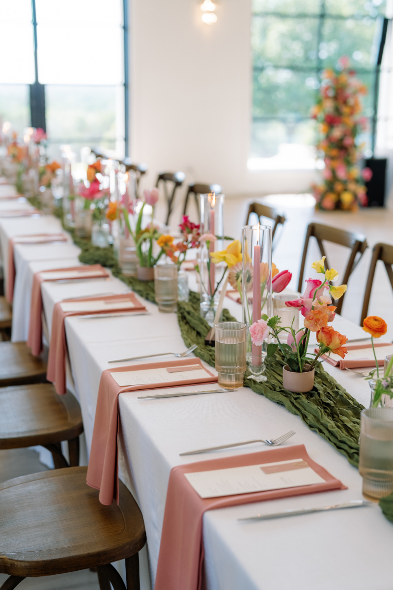 A long table set for a formal event with wooden chairs, white tablecloth, pink napkins, glassware, menus, candles, and colorful floral centerpieces arranged along the middle.