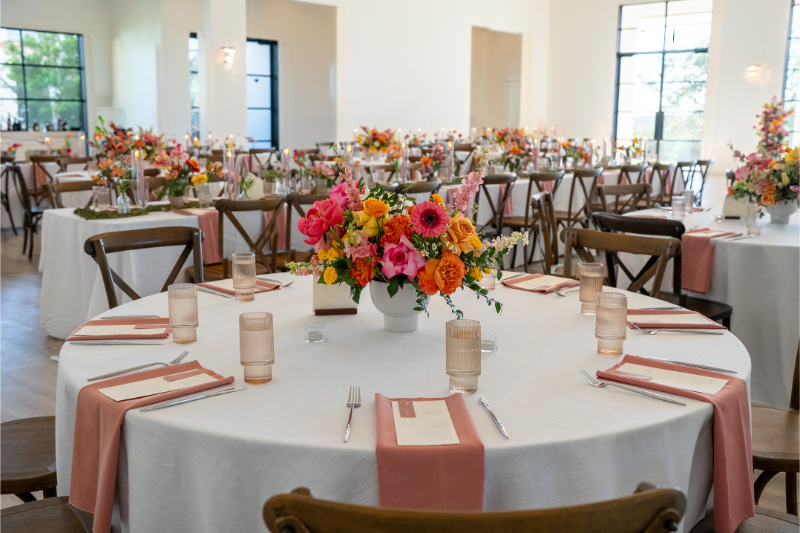 A round table set for an event with pink and orange floral centerpiece, place settings, and neatly folded napkins; similar tables fill a bright, airy room.
