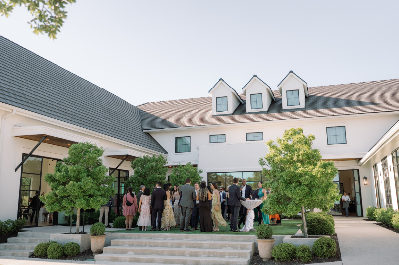 A group of people dressed in formal attire for cocktail hour at a wedding gather in courtyard of The Arlo Austin with large windows and greenery on a sunny day.