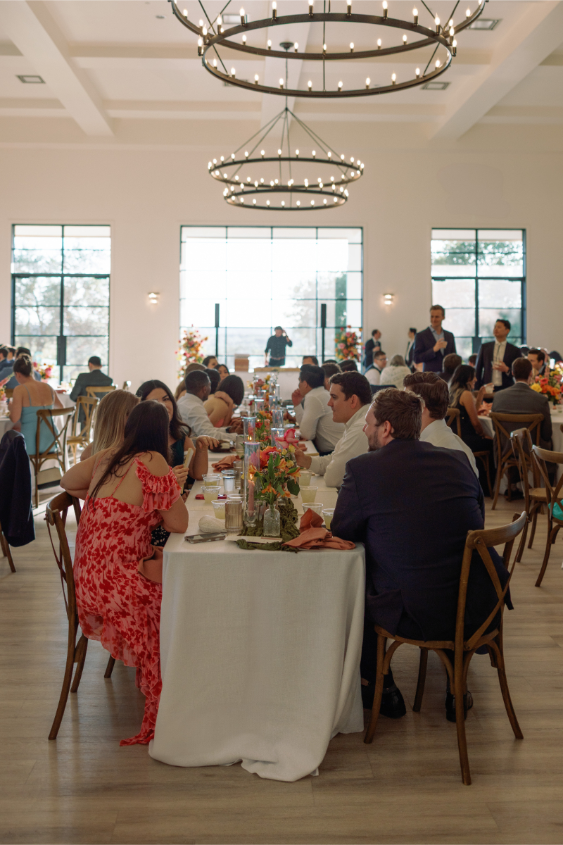 Guests sit at round tables in a well-lit event hall with large windows and chandeliers, decorated with floral centerpieces during a daytime gathering.