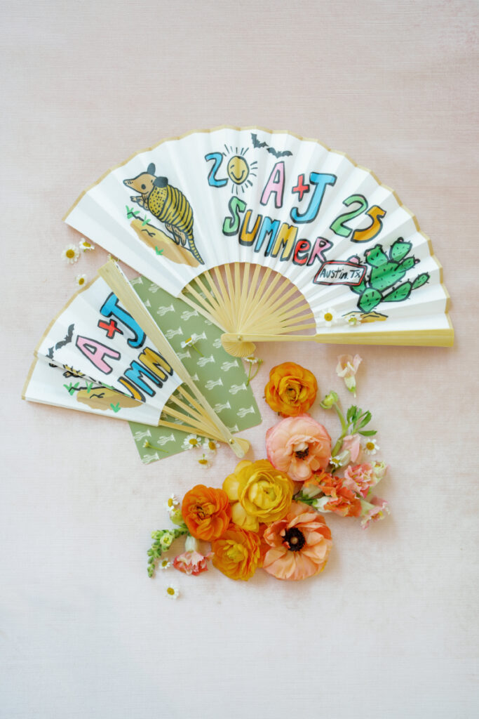 Two decorated paper fans with "A+J Summer 2025 Austin, TX" and colorful drawings, placed above orange and yellow flowers on a light pink surface.