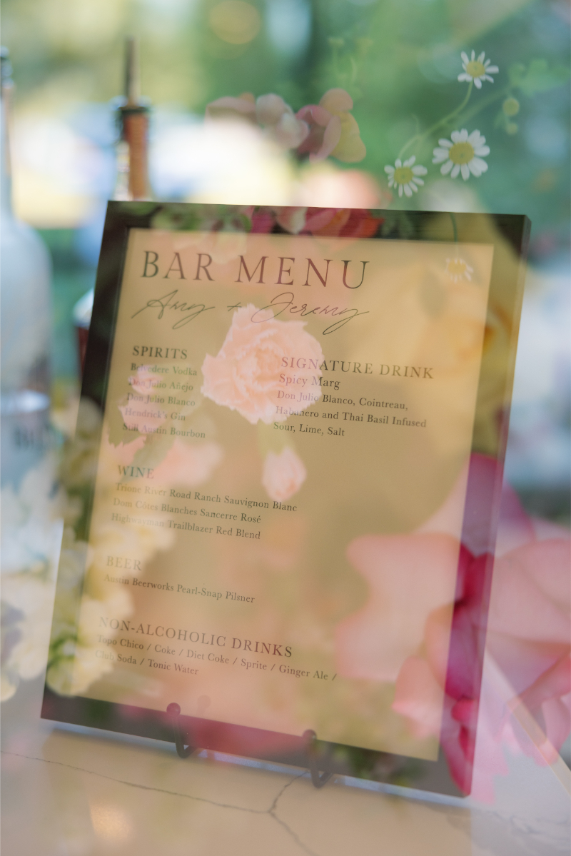 A bar menu featuring options for spirits, wine, beer, a signature drink called "Spicy Marg," and various non-alcoholic drinks, displayed with floral reflections on the glass.