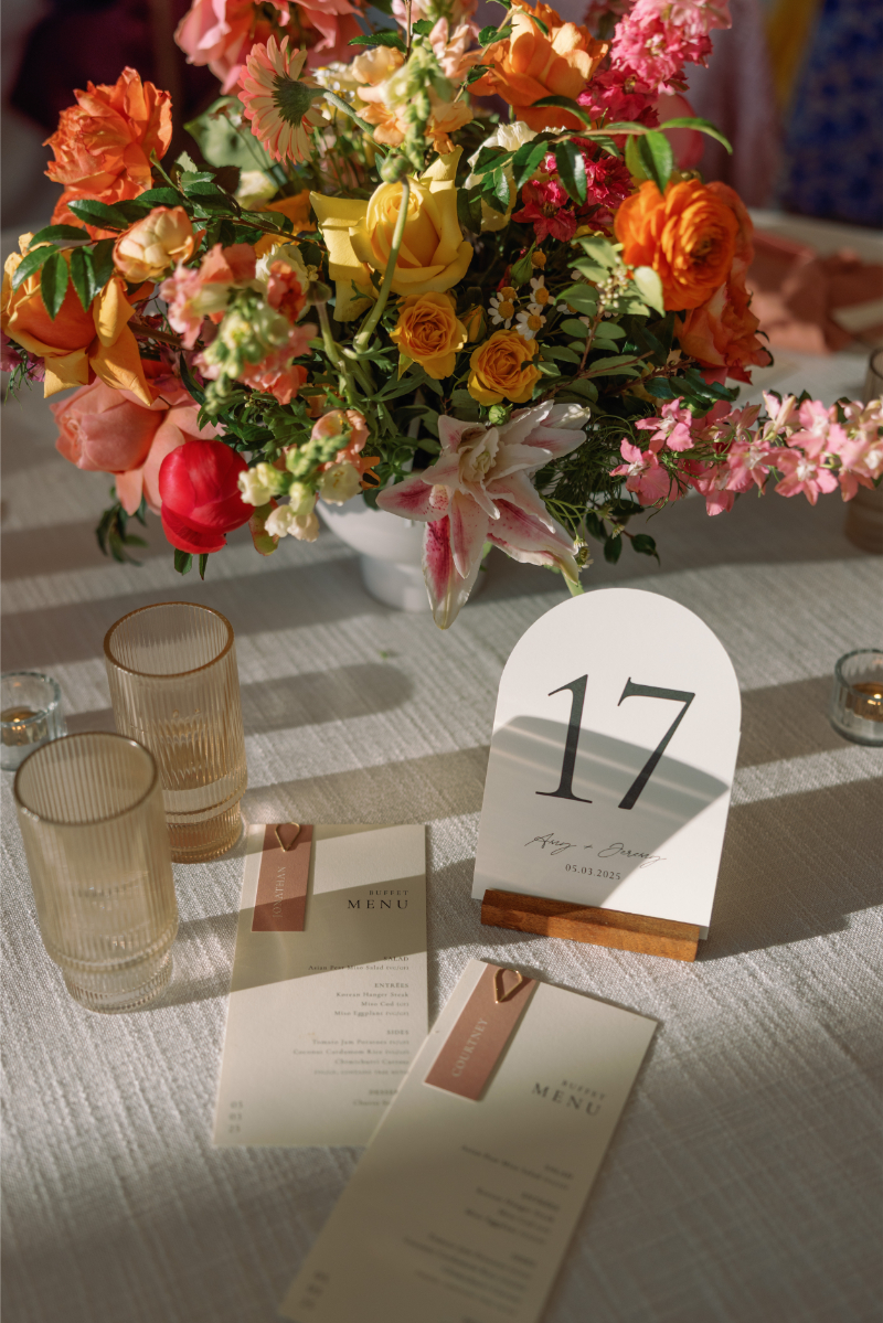 A table setting with a floral centerpiece, two beige glasses, two menus, and a table number card displaying "17" on a white tablecloth.