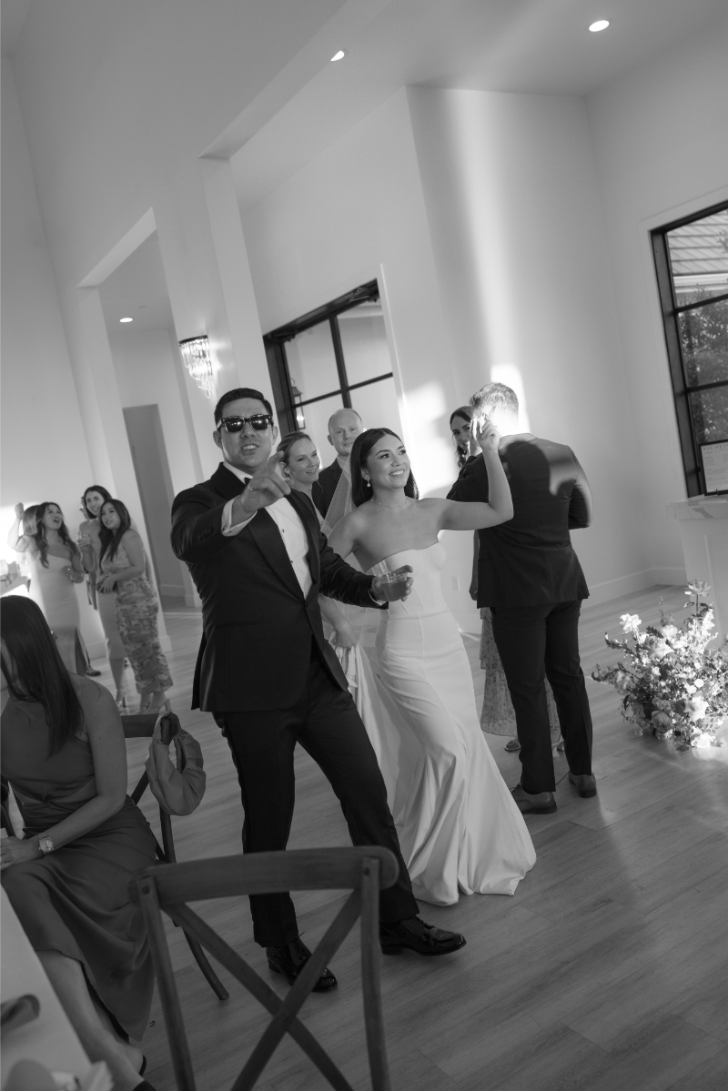 A bride and groom in formal attire walk and gesture joyfully at their wedding reception, surrounded by guests in a bright, modern venue.
