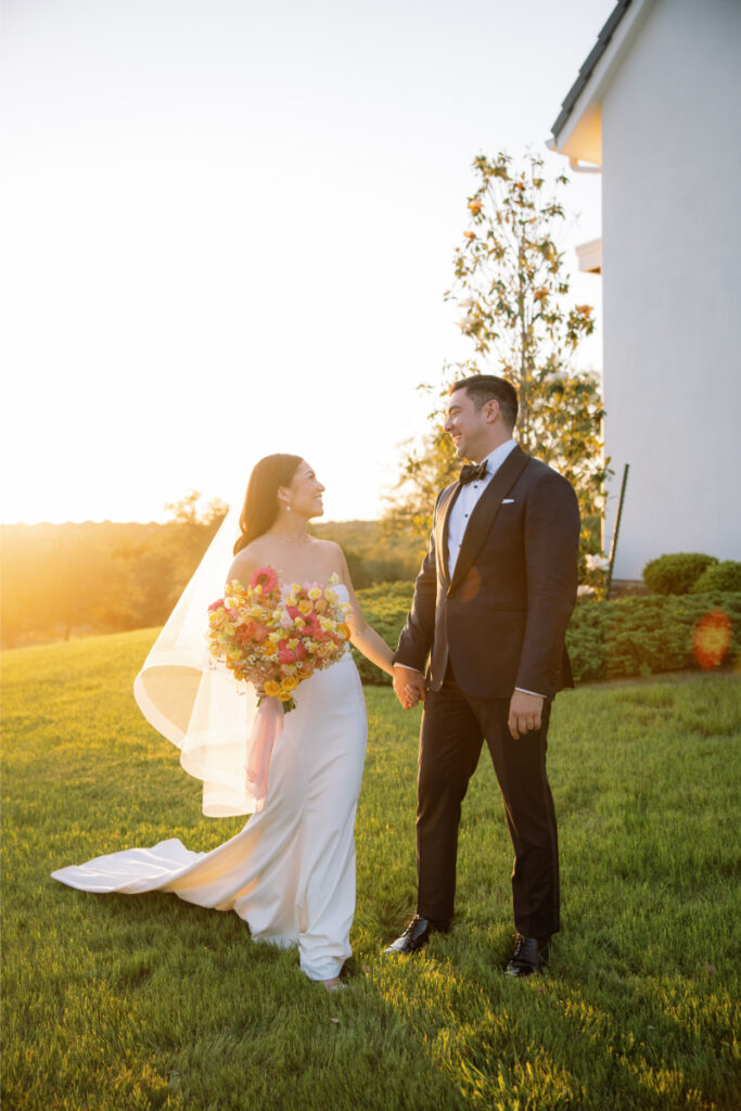 A bride in a white dress holding a bouquet and a groom in a black suit stand on grass outside, holding hands and smiling at each other at sunset.