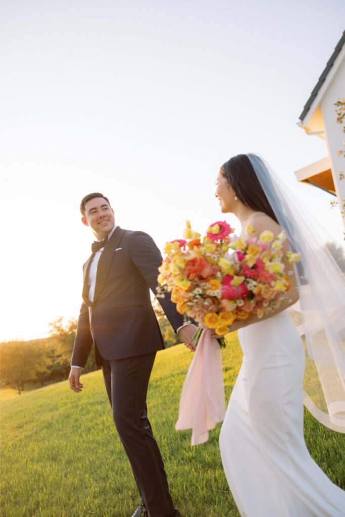 A bride in a white dress holding a colorful bouquet walks outdoors beside a groom in a dark suit, both smiling at each other with sunlight in the background.