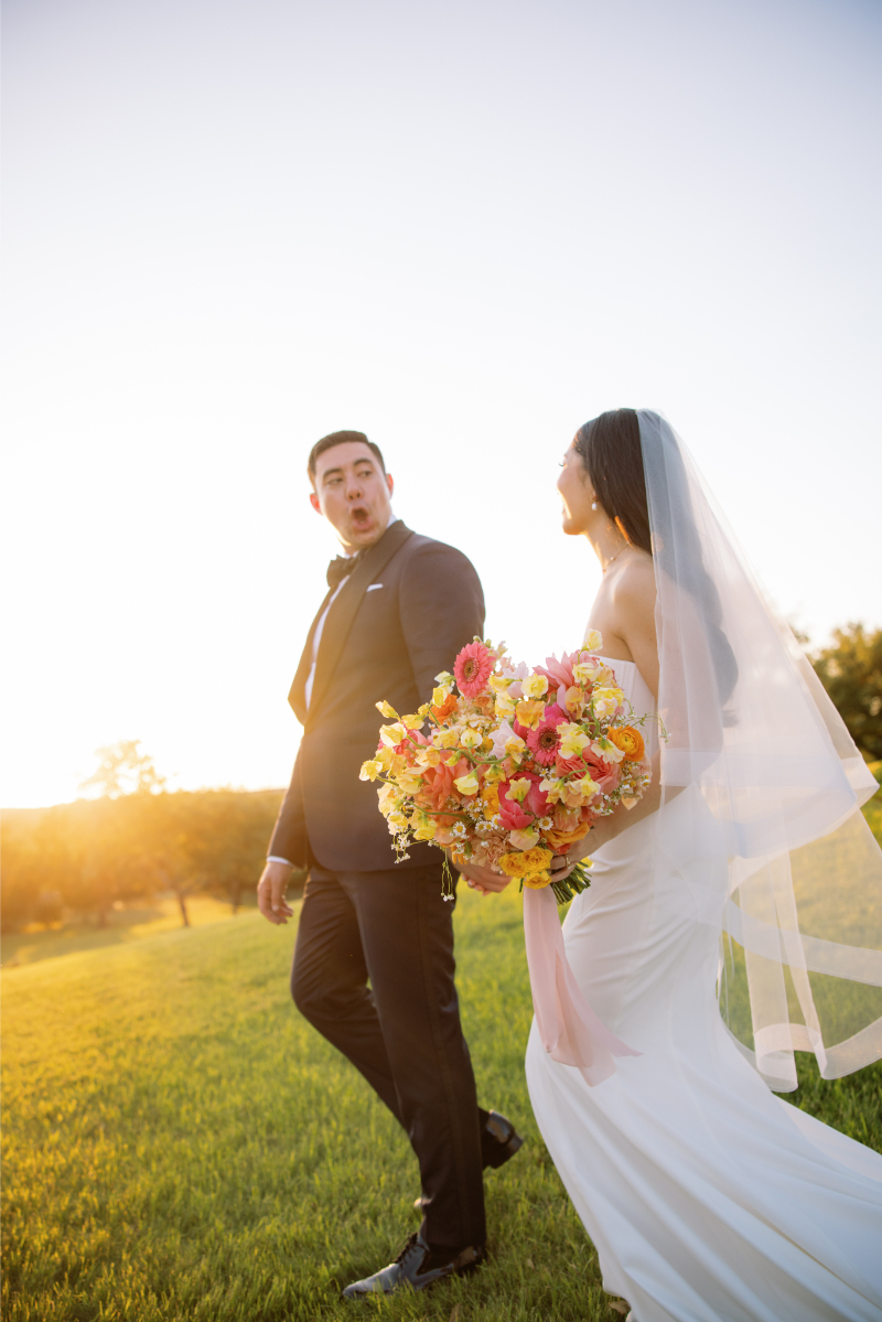 A bride in a white dress and veil holds a colorful bouquet while walking outdoors with a groom in a suit at sunset.