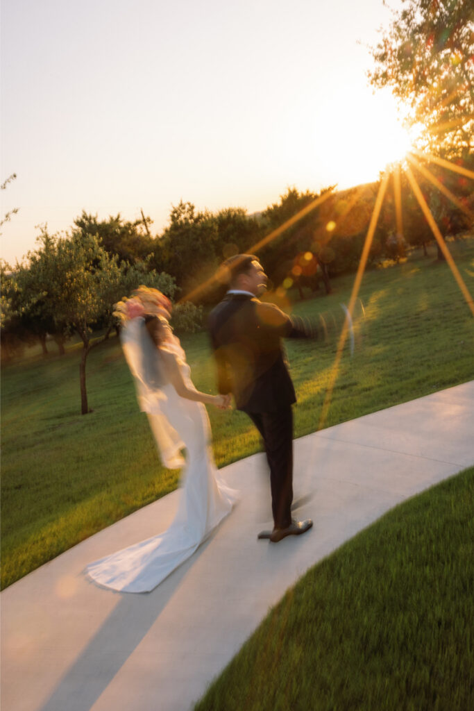A bride and groom walk hand in hand on a curved path through a grassy area at sunset, with sun rays shining in the background.