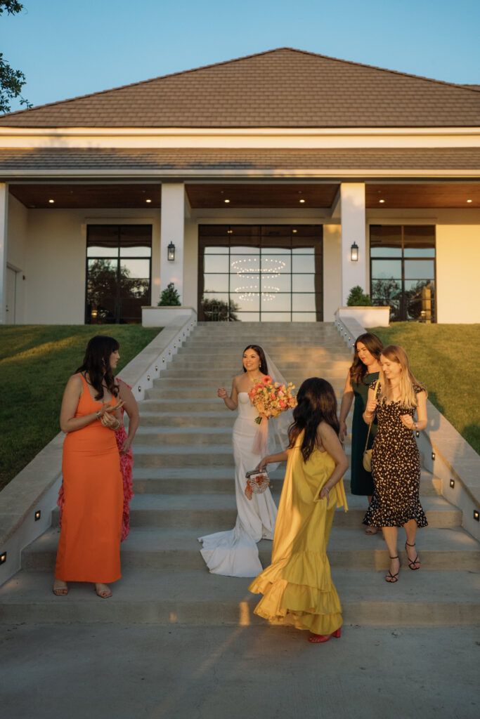 A bride in a white dress stands on outdoor steps with four women in colorful dresses, holding a bouquet and talking in front of a modern building at sunset.