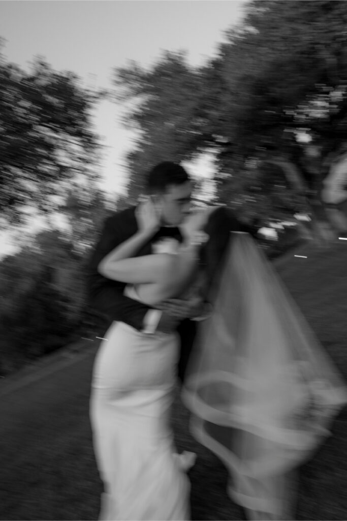 A bride and groom share a kiss outdoors; the image is black and white and noticeably blurred.