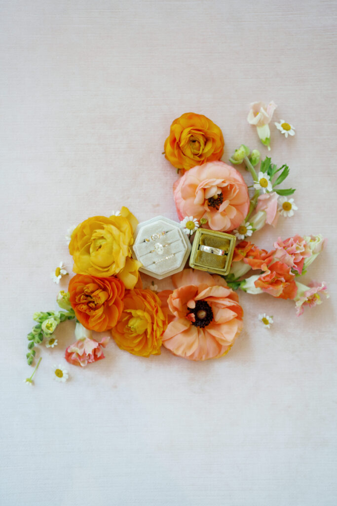 Two jewelry boxes with rings inside are surrounded by orange, peach, and yellow flowers on a light-colored surface.
