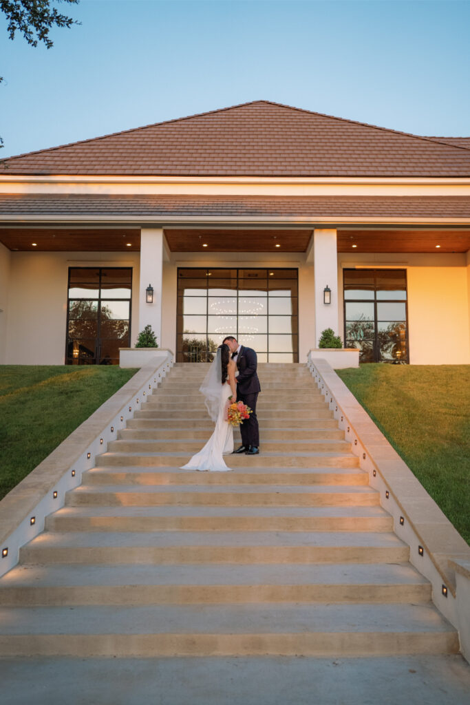 A bride and groom stand at the center of outdoor steps of The Arlo Austin, kissing in front of a modern building at sunset.
