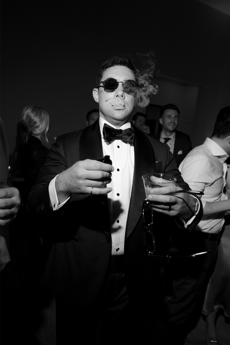 A man in a tuxedo and sunglasses holds a drink and a cigar, with smoke rising, at a crowded indoor event.