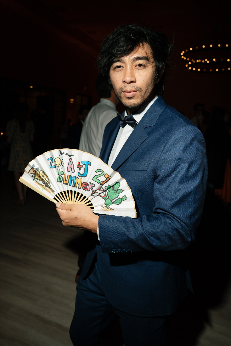 A man in a blue suit and bow tie holds a decorated fan with "2021 SUMMER" written on it at an indoor event.