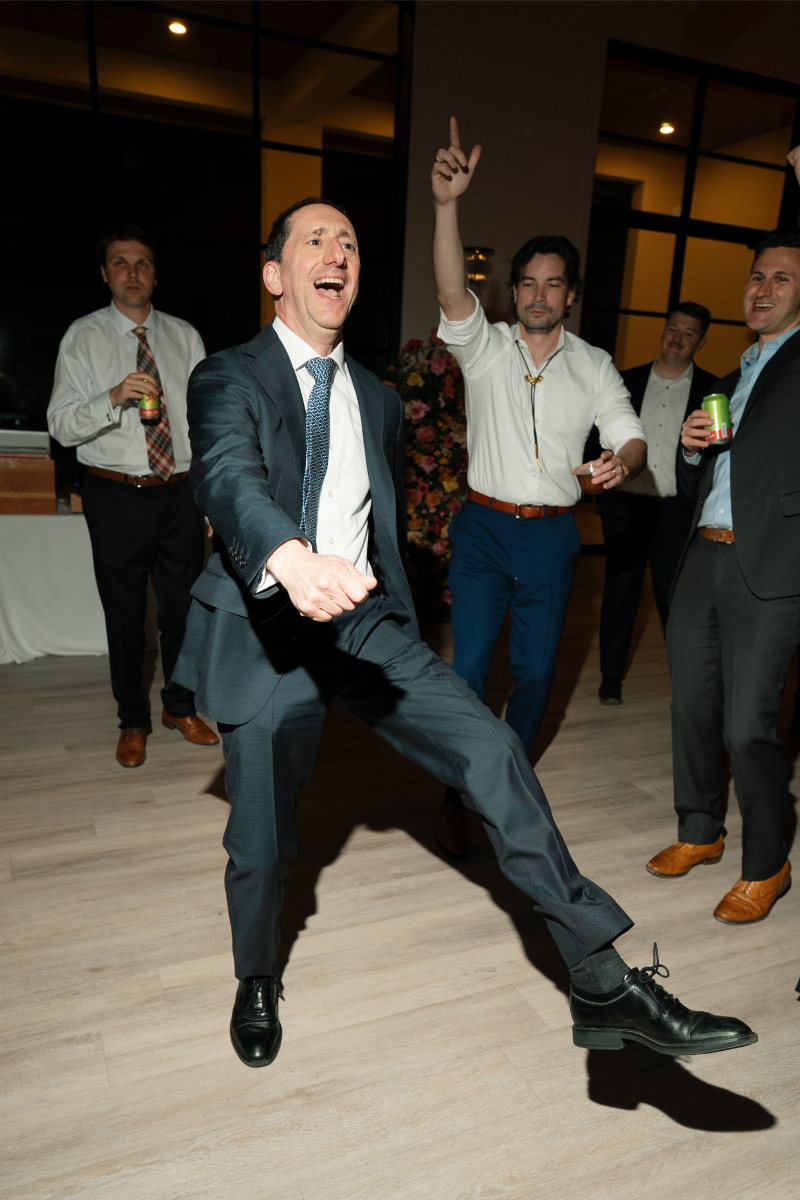 A man in a suit dances energetically on a wooden floor while several other men in dress clothes watch and cheer in the background.