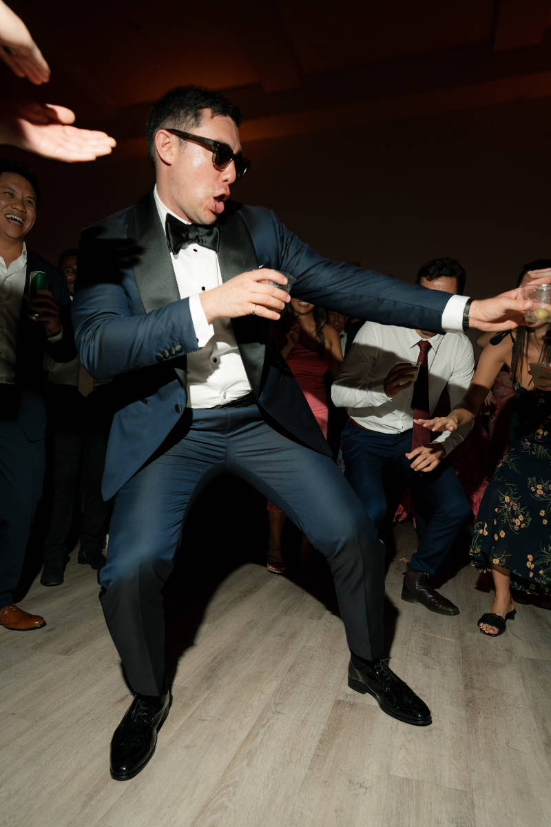 A man in a tuxedo and sunglasses dances energetically at a party, holding a drink, surrounded by other people on a dimly lit dance floor.
