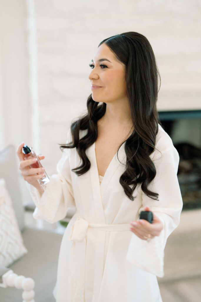 A woman with long dark hair, wearing a white robe, holds a bottle of perfume and sprays it on herself in a bright room.