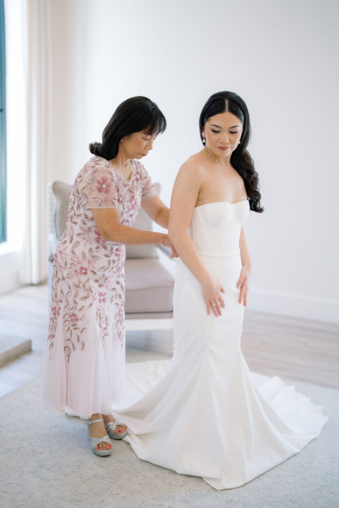 A woman in a floral dress helps another woman in a strapless white wedding gown adjust her dress in a bright room.