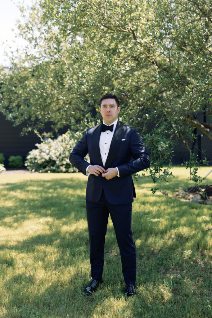 A man in a dark tuxedo stands on grass, buttoning his jacket, with trees and shrubs in the background on a sunny day.