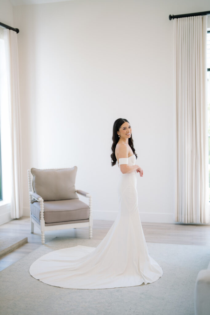 A woman in an off-the-shoulder white wedding gown stands inside The Arlo on a light rug, looking over her shoulder. Light streams in through large windows behind her.