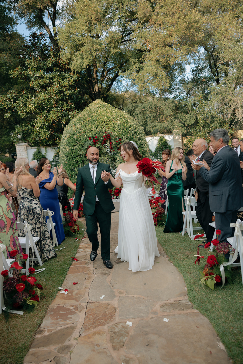 A bride and groom walk down an outdoor aisle lined with guests and red floral arrangements, holding hands and smiling after their wedding ceremony, great wedding design ideas for an outdoor ceremony.