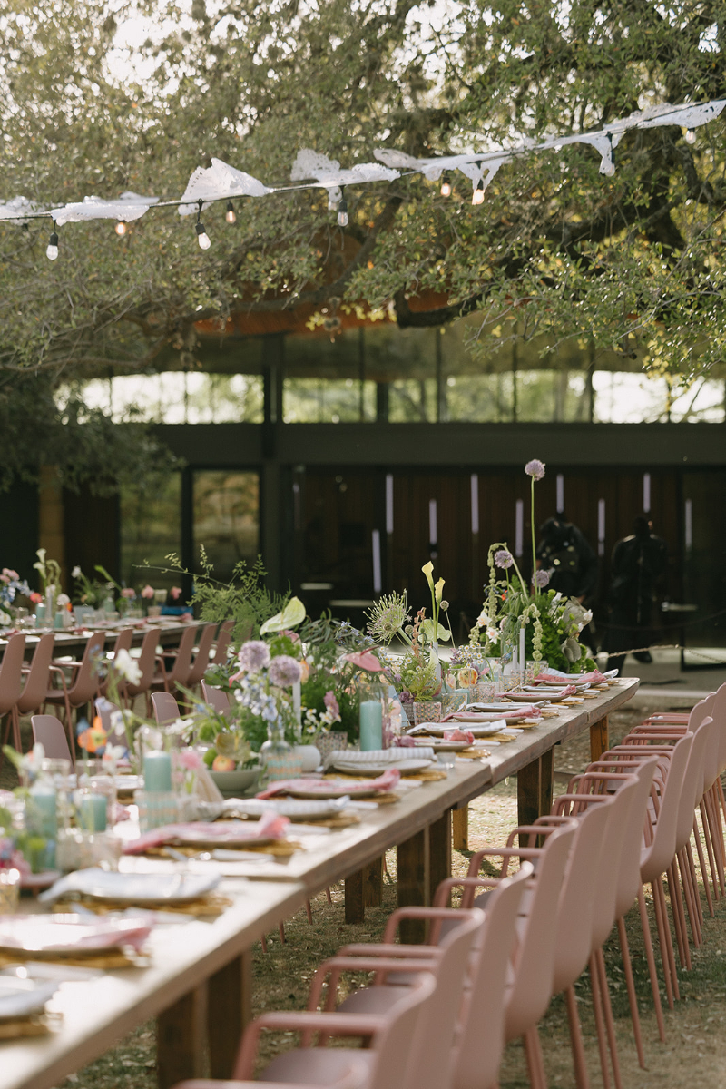 Wedding design ideas from a long outdoor table is set with floral centerpieces, plates, and glassware. Pink chairs line both sides, and string lights are hung above under trees.