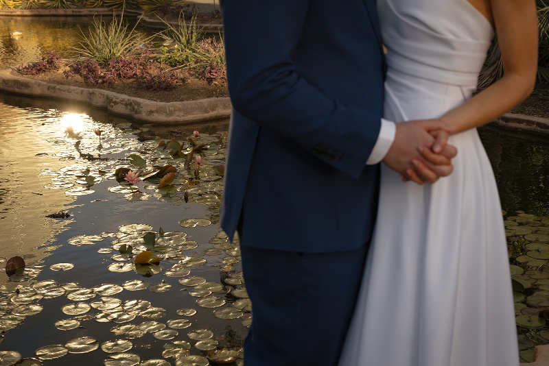 A couple dressed in formal attire, with the woman in a white dress and the man in a blue suit, stand holding hands near a pond with lily pads.