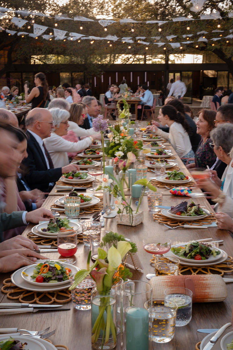 A large group of people sits around a long outdoor table set with plates of salad, drinks, and floral centerpieces at a gathering or celebration.